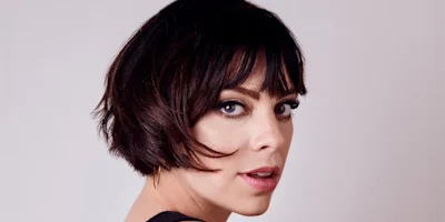 A woman with short dark hair and bangs looks over her shoulder toward the camera, against a plain light background.