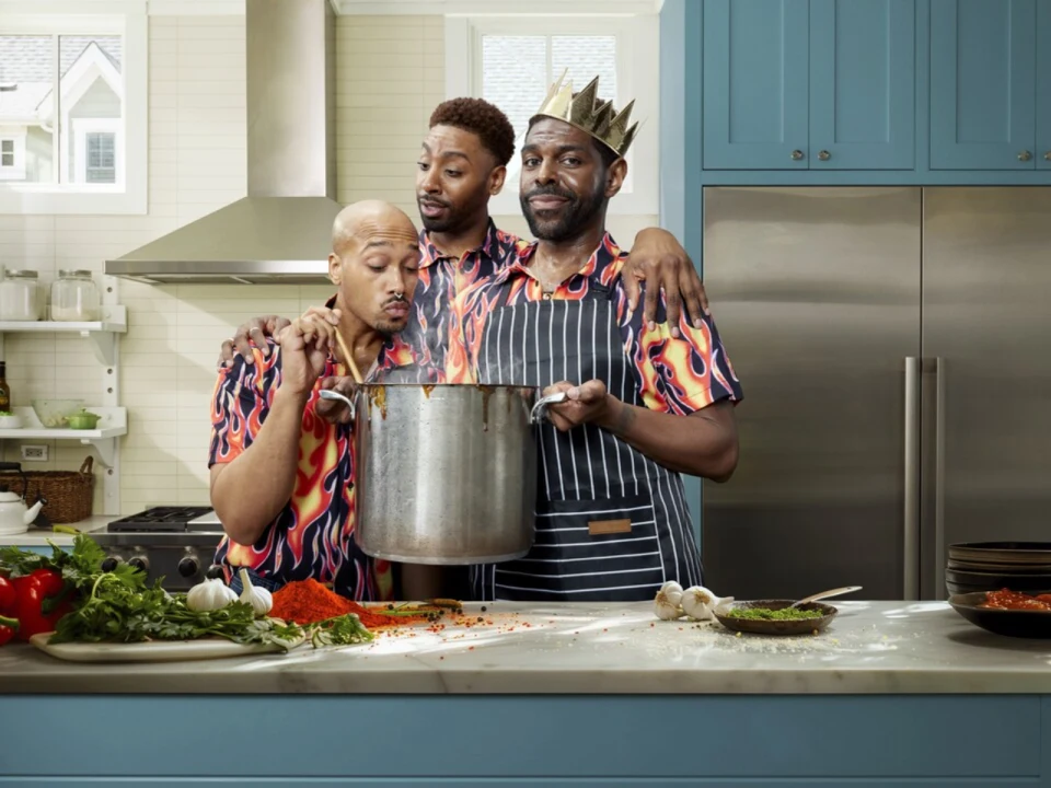 Three men in matching flame-patterned shirts are cooking in a kitchen. One man is stirring a pot, another is looking over his shoulder, and the third, wearing a paper crown, is smiling at the camera.