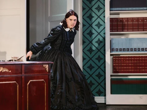 A person in a dark, Victorian-era outfit appears alarmed as they stand in a room with a wooden desk and a bookshelf filled with books.