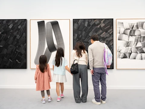 Four people, two adults and two children, stand closely together viewing abstract black and white artworks displayed on a gallery wall.