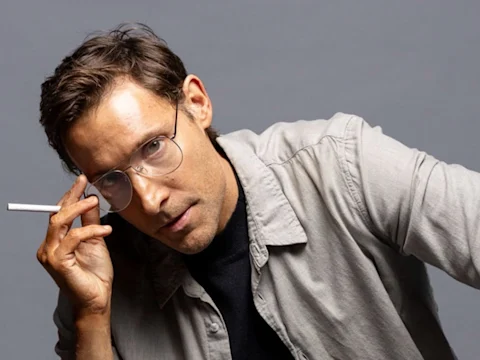 A man wearing glasses and a light gray shirt holds a cigarette near his face while leaning forward against a plain gray background.