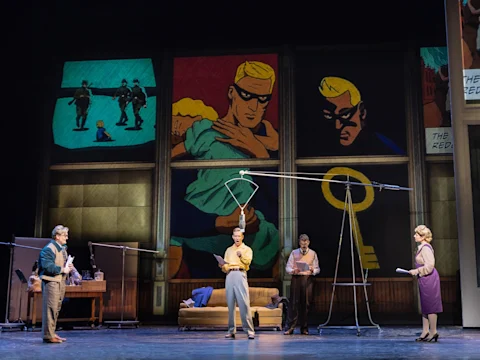 Four actors perform on a stage set resembling a living room, with a large comic book art backdrop depicting a blond superhero.