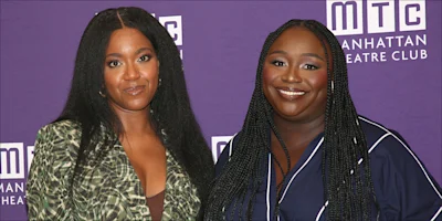 Two women stand together in front of a purple Manhattan Theatre Club backdrop, one wearing a green patterned blazer, the other a navy blue outfit with white trim.