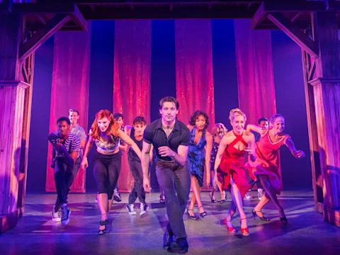 A group of musical theater performers in dance attire moving toward the audience on a stage with red vertical curtains.