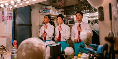Three men in dress shirts and ties stand in front of a well-lit dressing room mirror, adjusting their ties, with wigs and styling tools visible on the counter.