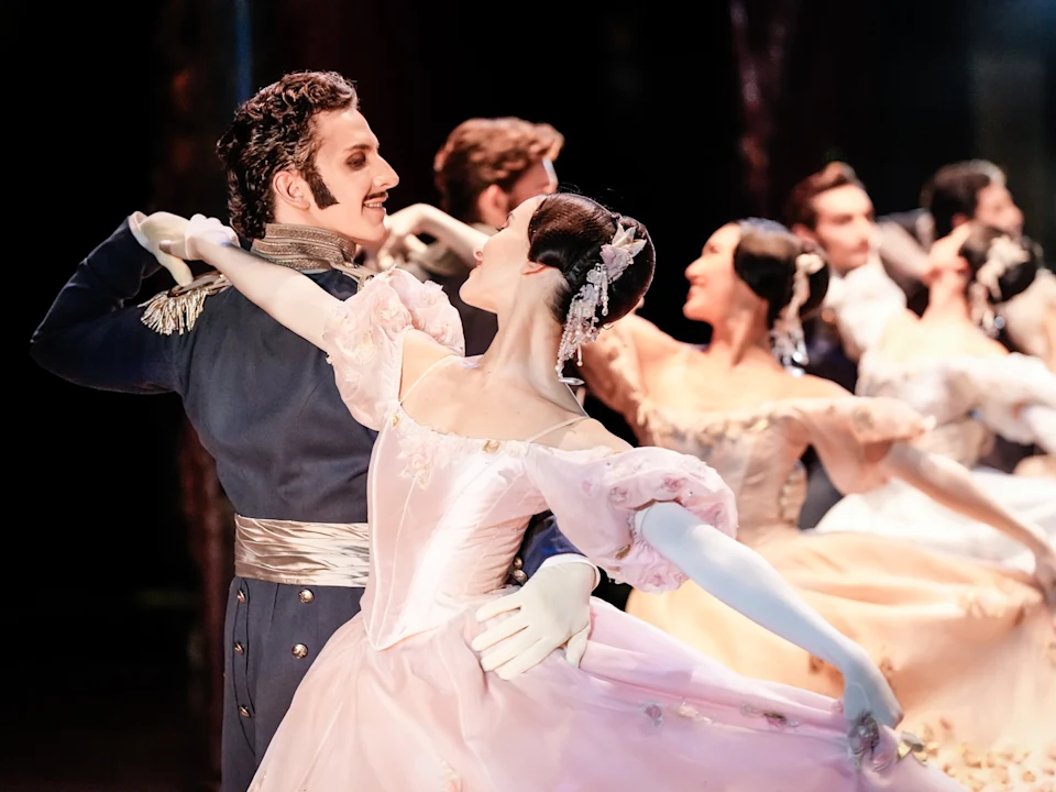 A group of ballet dancers in formal costumes perform a synchronized dance; a man and woman in the foreground hold an elegant pose.