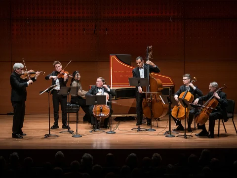 A chamber music group performs on stage with various string instruments, including violins, a cello, a double bass, and a harpsichord. The musicians are formally dressed and engaged in their performance.