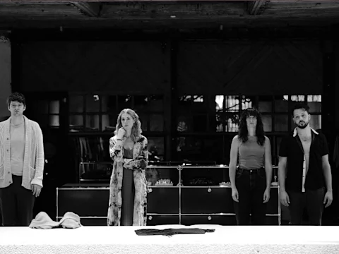 Four people stand side by side indoors, facing forward with neutral expressions. Some items, including hats, are placed on a surface in front of them. The background appears dark and industrial.