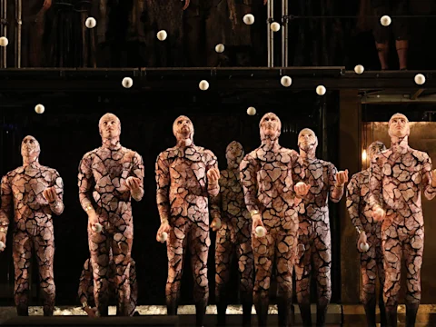 Production photo of Akhnaten in Los Angeles, showing seven performers in patterned bodysuits juggle white balls in unison on a stage, with more balls suspended mid-air above them.