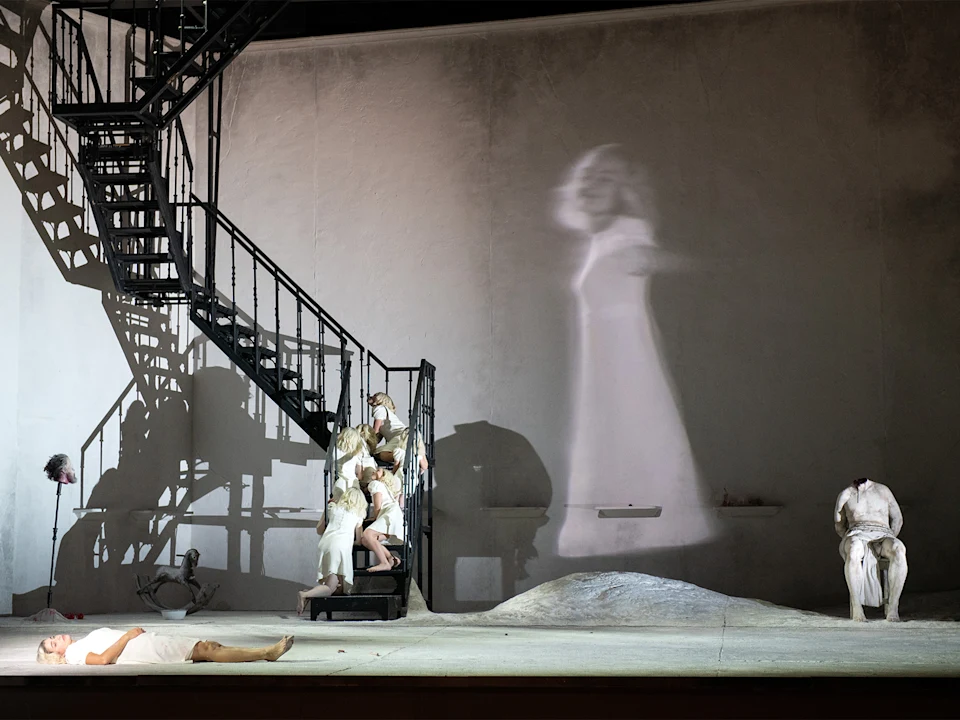 Theater scene with actors in white costumes on a minimalist set. A shadow of a figure in a dress is projected on a wall beside a staircase.