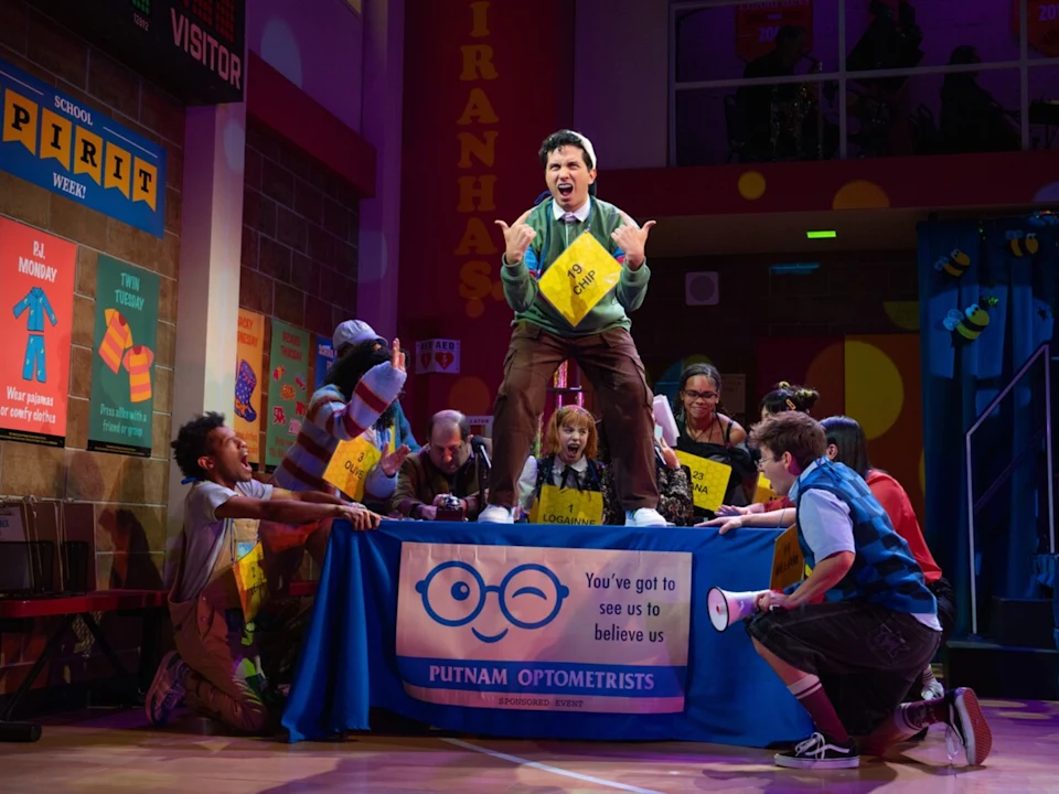 A group of actors on stage perform a lively scene around a table with a "Putnam Optometrists" banner; one actor stands on the table holding a yellow folder.