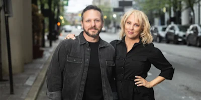 Two people stand side by side on a city street, both wearing dark button-up shirts. One person has their arm around the other’s shoulder. Cars and trees line the background.
