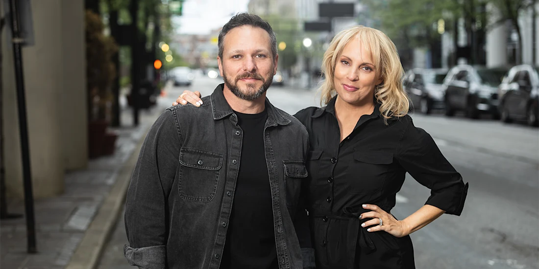 Two people stand side by side on a city street, both wearing dark button-up shirts. One person has their arm around the other’s shoulder. Cars and trees line the background.