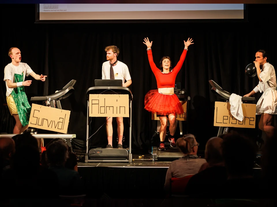 People on stage performing on treadmills with signs labeled "Survival," "Admin," and "Leisure," dressed in various costumes. A person raises their arms enthusiastically. Audience visible in foreground.