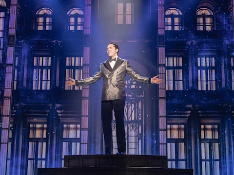 A person in a patterned suit stands on a raised platform with arms outstretched in front of a grand, illuminated building backdrop.