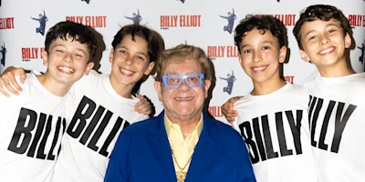A man in blue glasses and a blue suit stands with four smiling boys wearing "BILLY" T-shirts in front of a "Billy Elliot" backdrop.
