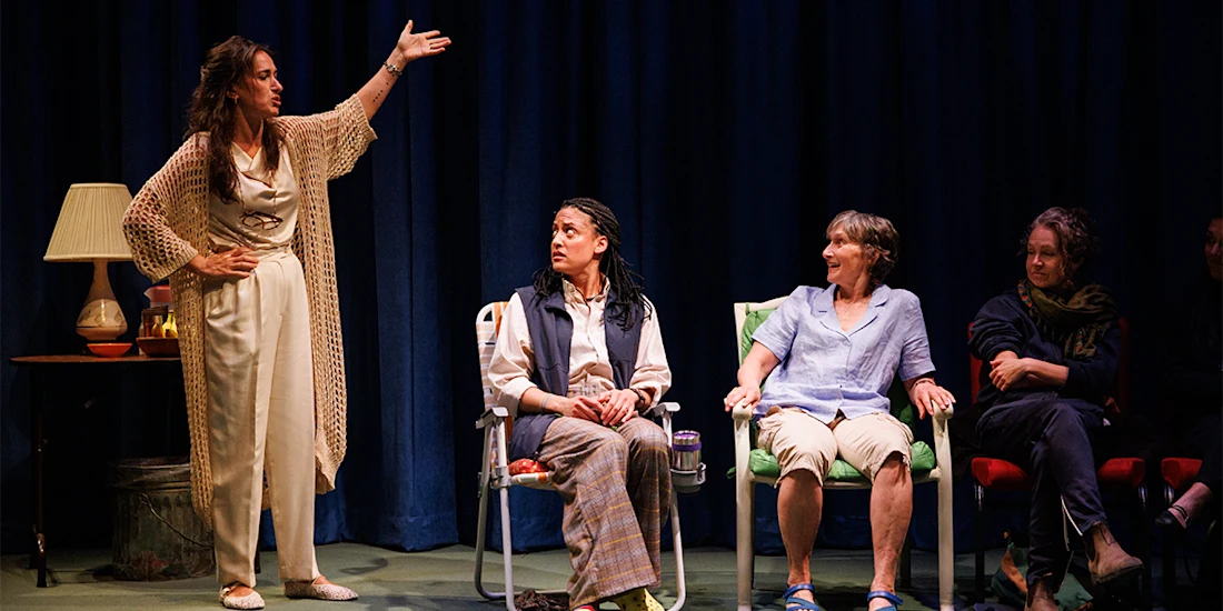 Four women are seated on stage with one woman standing and gesturing while the others look at her; a lamp and table are to the side, and a blue curtain is in the background.