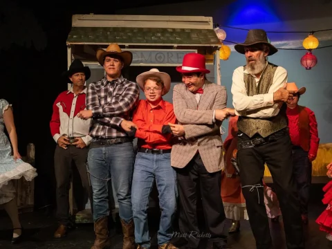 Group of people in cowboy and western attire perform a synchronized dance on stage. Some wear hats, with a costume house backdrop and decorative lanterns above.