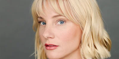 A woman with short blonde hair and blue eyes looks at the camera against a plain gray background.