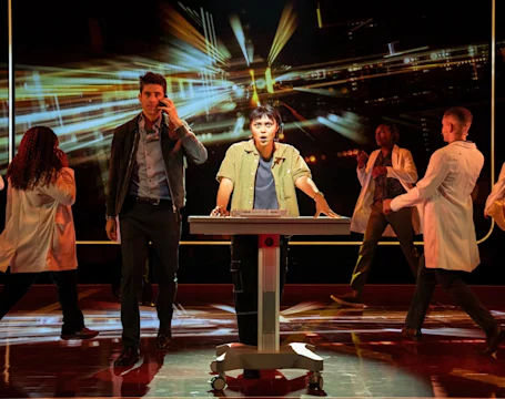 A cast member at a central podium looks surprised while surrounded by others in a stage performance against a background of motion-blurred lights.