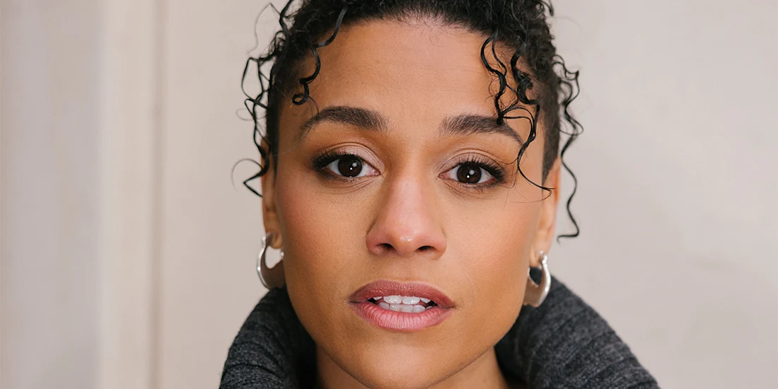 A person with curly hair and hoop earrings looks directly at the camera against a plain background.