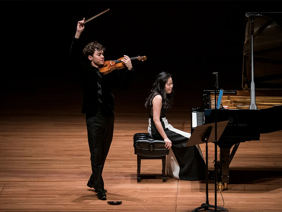 A violinist stands with his bow raised while a pianist sits at a grand piano on a wooden stage during a performance.