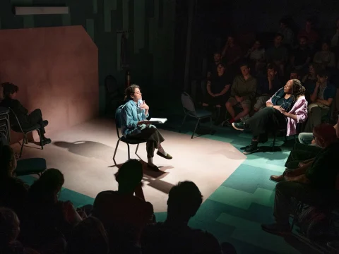 Two people seated on stage engaging in a conversation, surrounded by an audience in a dimly lit room.