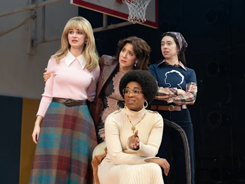 Four women stand on a gymnasium floor, all looking in the same direction with expressions of curiosity and concern. A basketball hoop is visible in the background.