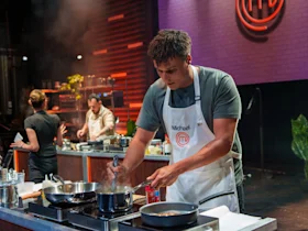 A contestant in a white apron labeled "Michael" cooks at a stovetop during a televised cooking competition, with two other participants in the background.