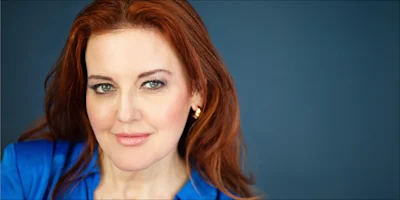 Woman with long red hair wearing a blue top and gold earrings, looking at the camera against a plain dark blue background.