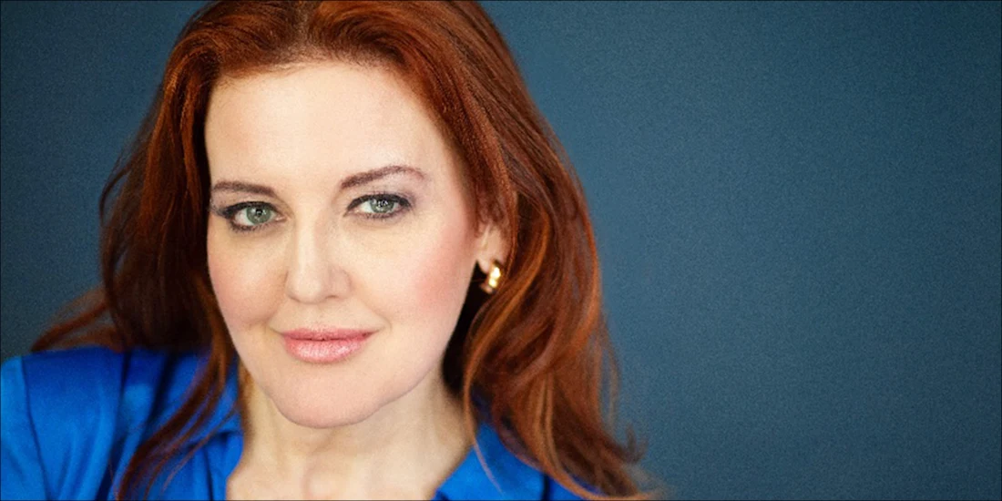 Woman with long red hair wearing a blue top and gold earrings, looking at the camera against a plain dark blue background.