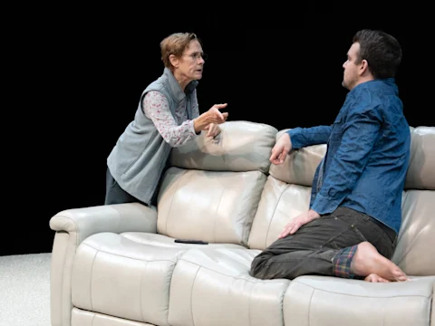 An older woman stands and gestures while talking to a seated man on a beige couch; both appear engaged in a serious conversation.