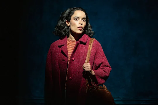 A woman with dark hair, wearing a red coat and carrying a brown bag, stands in front of a dark blue textured background, looking slightly to the side.