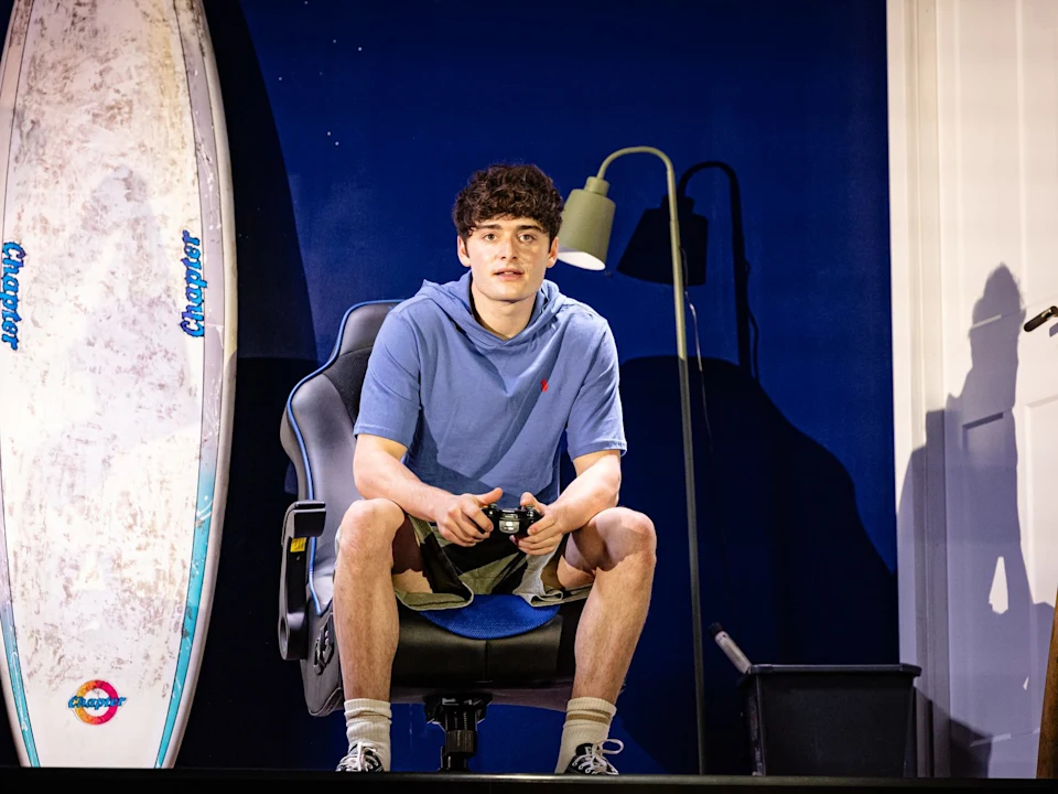 A teenage boy sits in a gaming chair holding a games controller, a surfboard propped beside him in his bedroom.