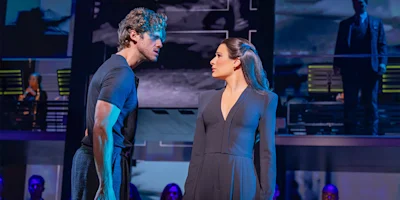 A man and a woman stand facing each other on stage, surrounded by blue lighting and other performers in the background.
