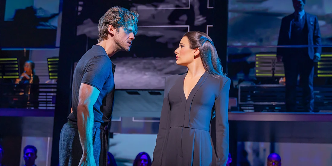 A man and a woman stand facing each other on stage, surrounded by blue lighting and other performers in the background.
