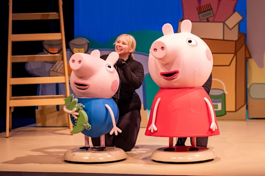 A performer operates large Peppa Pig and George Pig character puppets on a stage with cartoon-style props in the background.