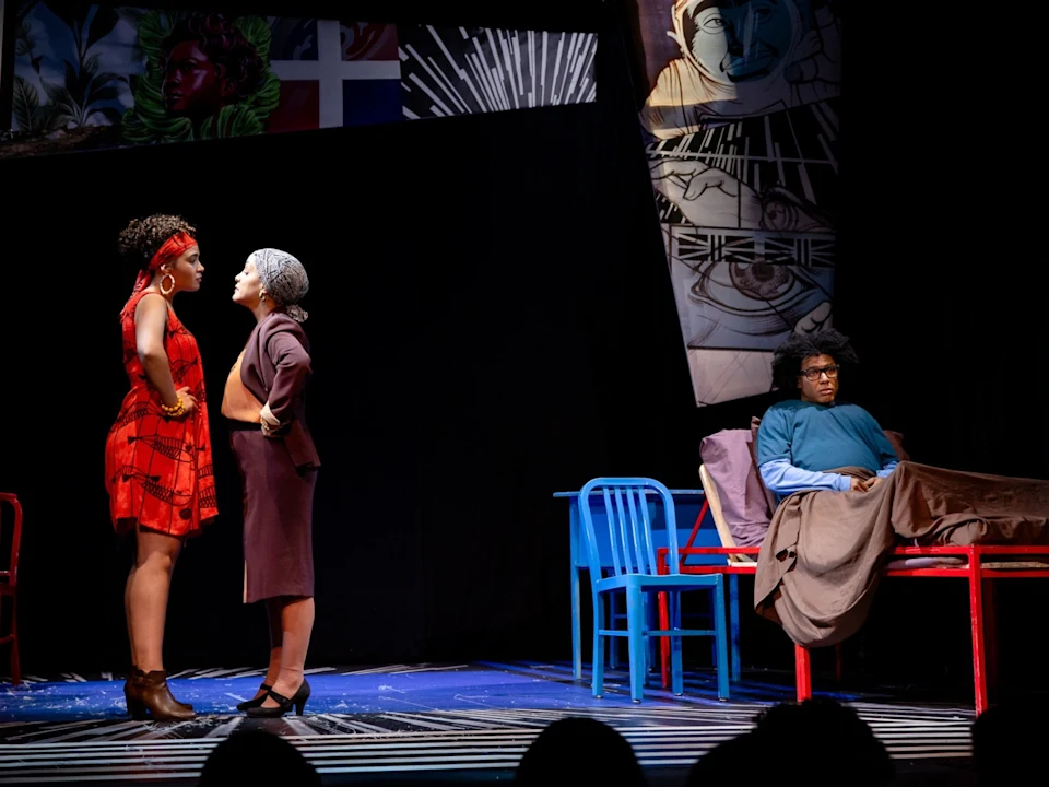 Two women stand facing each other in conversation while a person sits on a bed covered with a blanket, observing them; colorful chairs and murals are visible in the background.