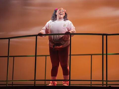 A person in a pink sweater and red leggings stands smiling on a stage with an orange lit background, holding a green railing.