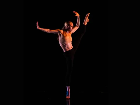 A dancer performs a high arabesque in a dark setting, wearing a light-colored top and dark pants, with one leg raised high and arms extended.