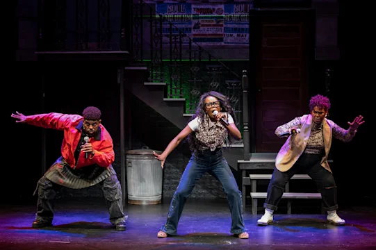 Three performers on stage strike dynamic poses during a musical number, with urban-themed set pieces in the background.