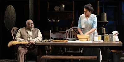 A man sits at a table holding a broom while a woman stands beside him mixing ingredients in a bowl in a rustic kitchen setting.