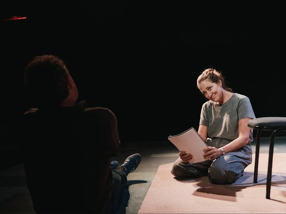 A person sits on the floor holding papers, looking at another person who is seated nearby under dim lighting.