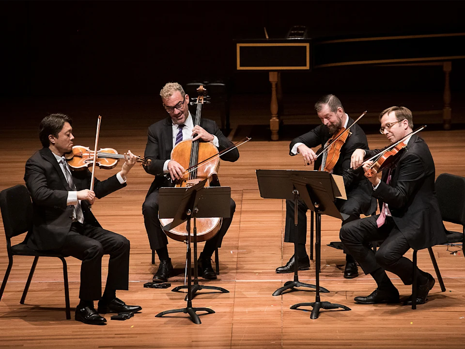 Four musicians in suits perform on stage, playing violin, cello, and viola, each seated with a music stand in front of them.