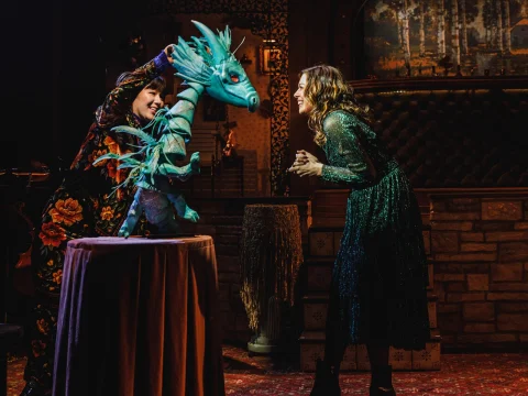 A person manipulates a blue dragon puppet as another person reacts nearby in a dimly lit room with medieval decor.