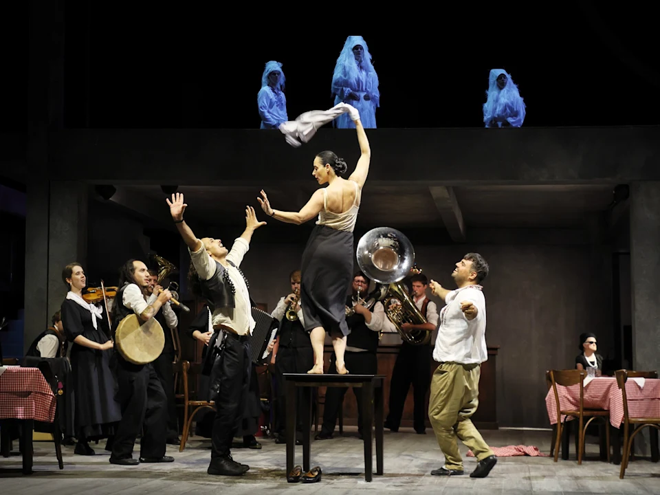 A woman stands on a table waving a cloth as musicians and actors perform around her; three blue-lit figures watch from above.