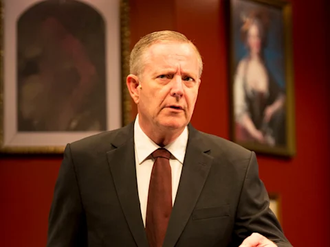 A man in a suit and tie stands indoors, looking serious. Two framed portraits hang on the red wall behind him.