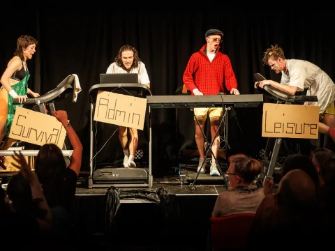 Four people are on treadmills on stage, labeled "Survival," "Admin," and "Leisure," with an audience watching.