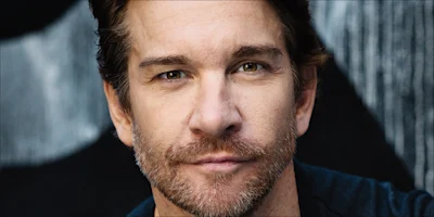 A middle-aged man with short brown hair and light stubble looks directly at the camera with a neutral expression. The background is dark and blurred.