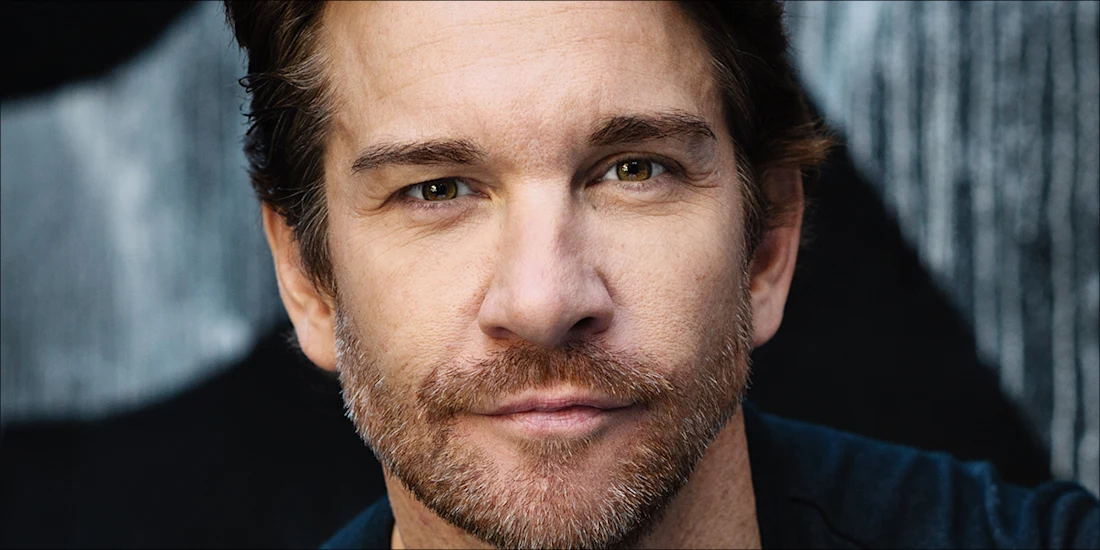 A middle-aged man with short brown hair and light stubble looks directly at the camera with a neutral expression. The background is dark and blurred.